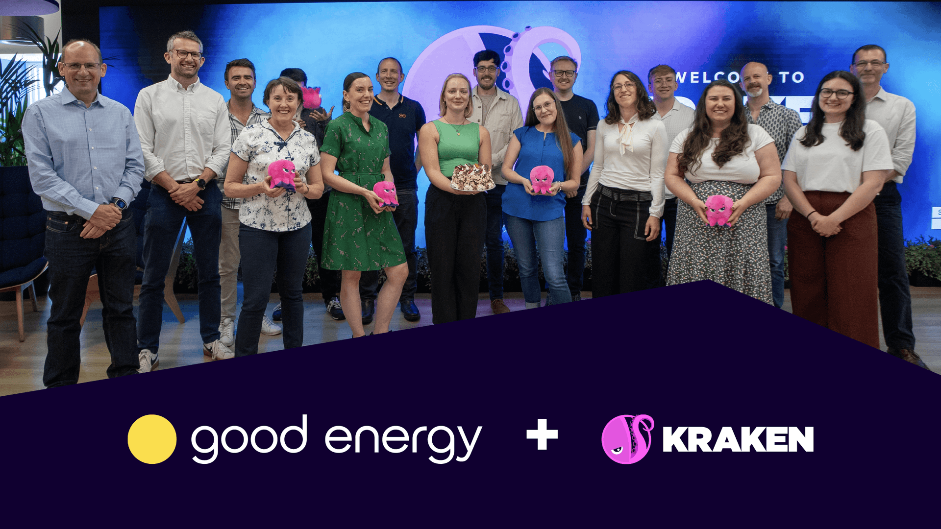 Two rows of people from the Good Energy and Kraken teams, some holding Kraken plushies, in the Kraken office. Behind, there's a screen with the Kraken logo. 